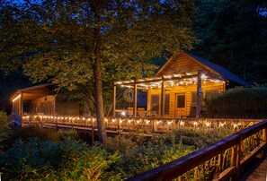 Uma cabana de madeira com varanda e luzes de cordão, rodeada por árvores e um deck de madeira.