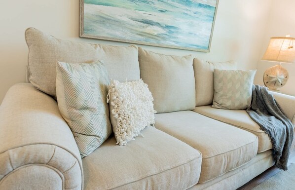Un sofá seccional color beige con varios cojines, un cuadro colgado en la pared, una mesita de noche con una lámpara y una concha decorativa.