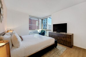 A modern bedroom with a large bed, a TV on a wooden dresser, and a view of buildings through a large window.