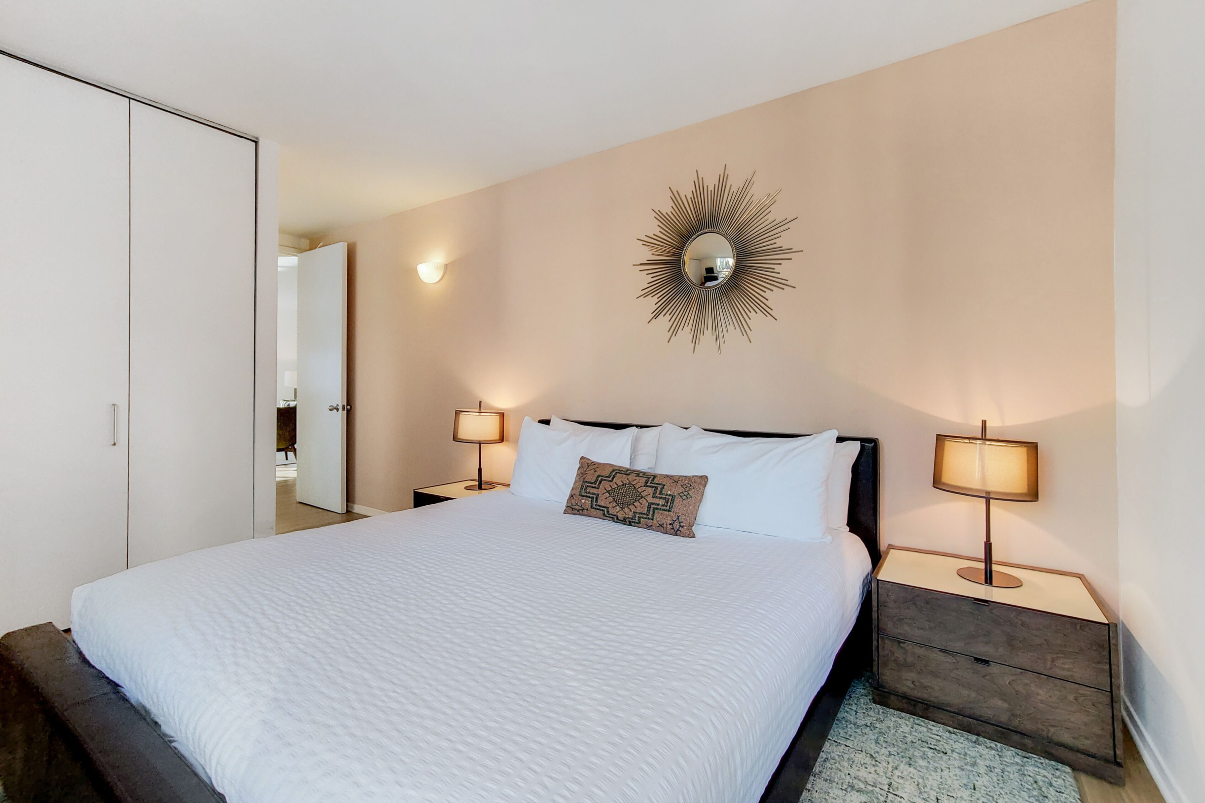 Cama bem arrumada com roupa de cama branca, um espelho decorativo na parede e duas luminárias de cabeceira.