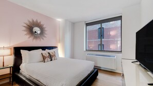 Un dormitorio moderno con una cama grande, un televisor, una ventana que ofrece vistas a un edificio de ladrillo y un espejo de pared original.