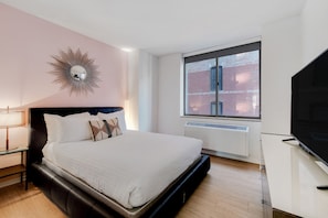 Ein modernes Schlafzimmer mit einem großen Bett, einem Fernseher, einem Fenster mit Blick auf ein Backsteingebäude und einem außergewöhnlichen Wandspiegel.