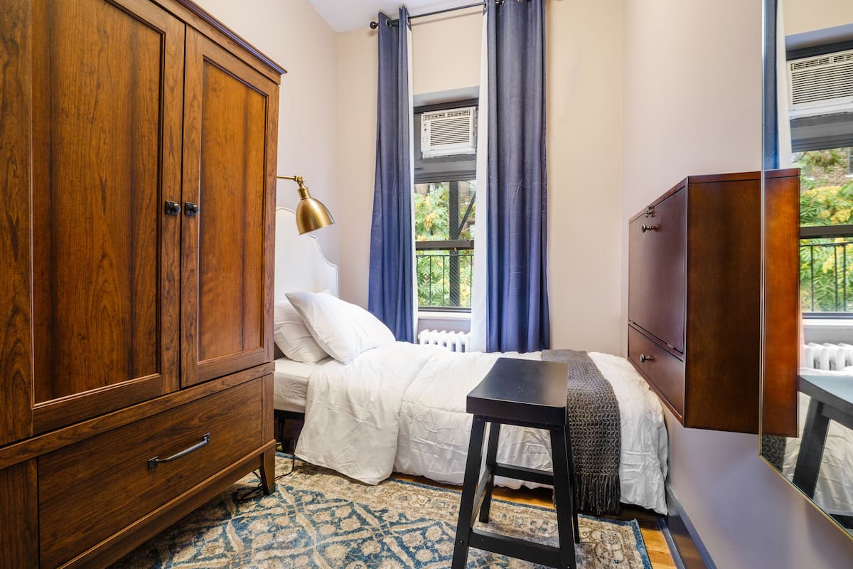 A bedroom with a wooden wardrobe, a bed with white bedding, a small table, and a window with curtains.