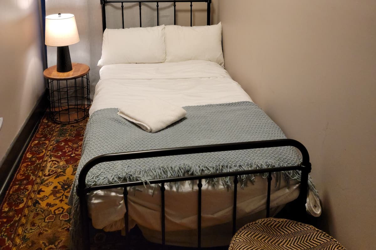 A small, neatly made bed with a metal frame, a bedside lamp, and a patterned rug.