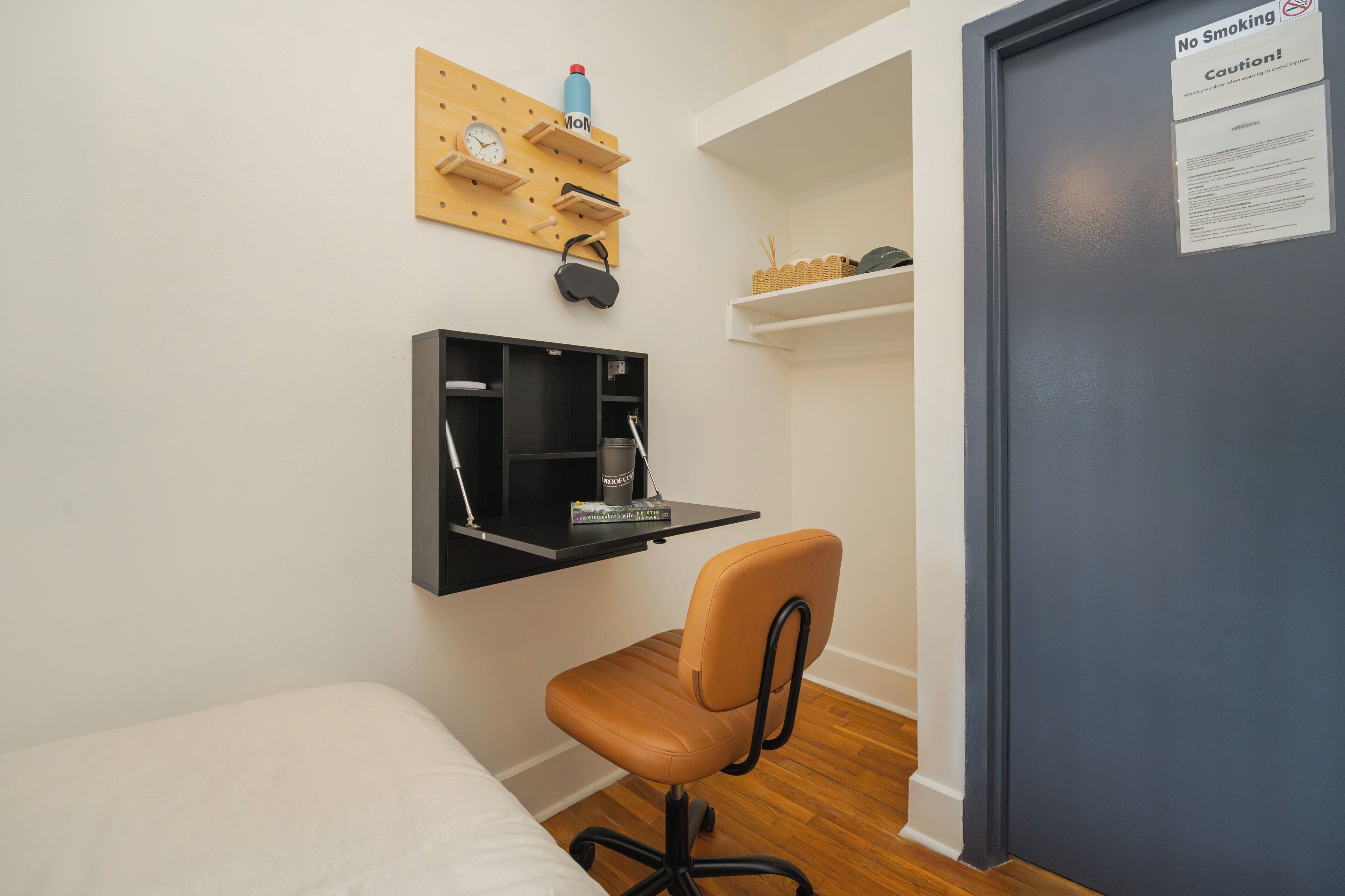 Una habitación pequeña y funcional con cama, escritorio y silla, estante en la pared con un reloj y una puerta con un letrero de 'No Fumar'.