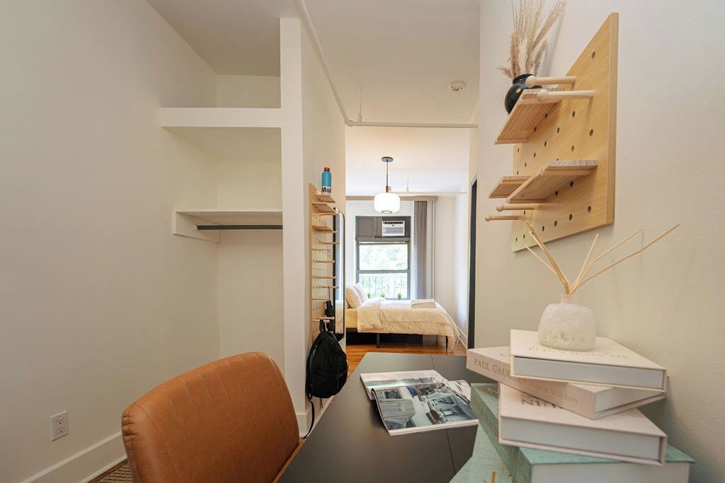 A modern bedroom with a bed, a desk with a chair, a wall-mounted shelf with books, and a hanging light fixture.
