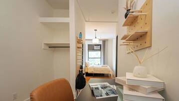 A modern bedroom with a bed, a desk with a chair, a wall-mounted shelf with books, and a hanging light fixture.