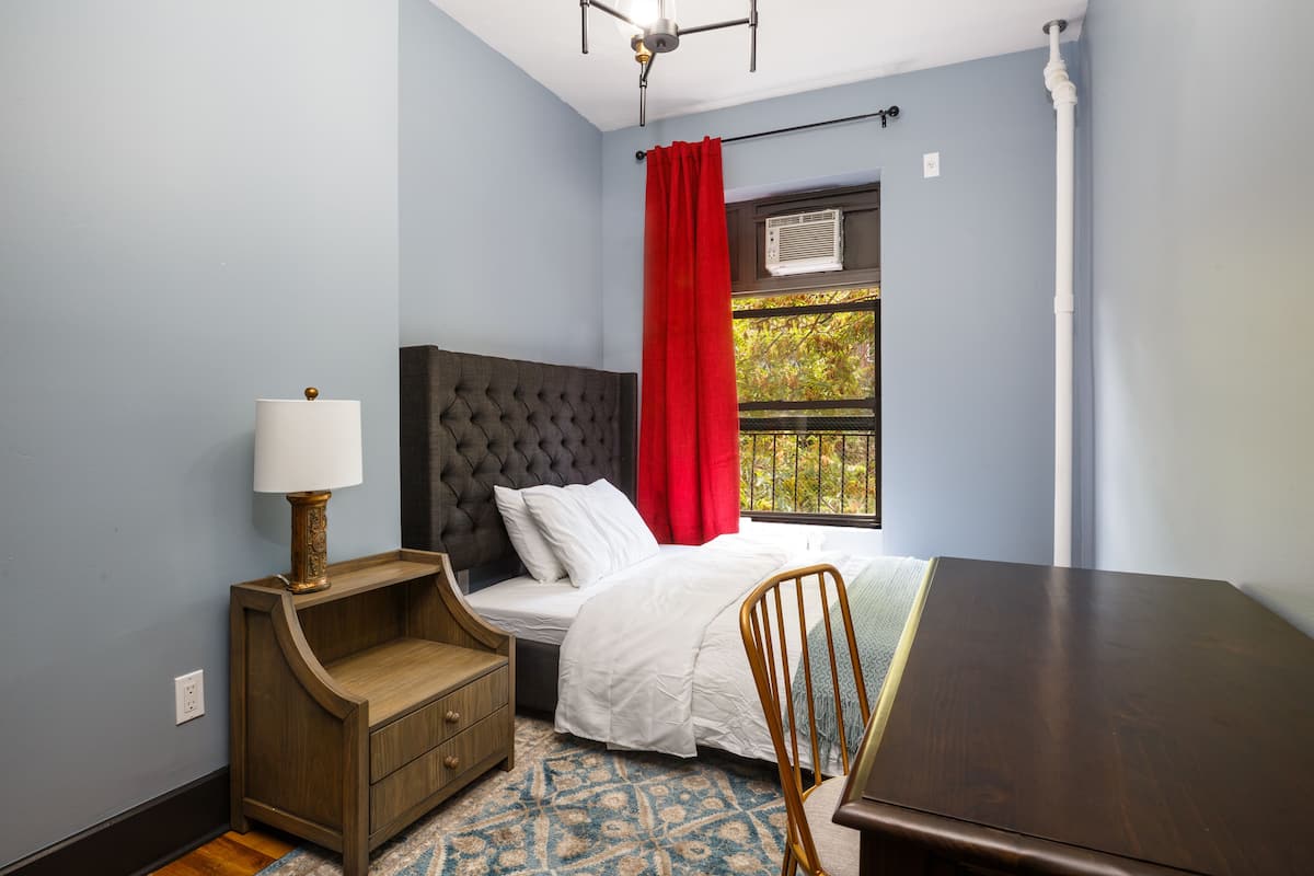 A bedroom with a bed, a nightstand, a chair, a table, and a window with curtains.