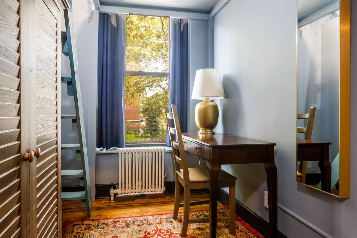A room with a wooden desk, a chair, a lamp, a radiator, and a window with curtains.