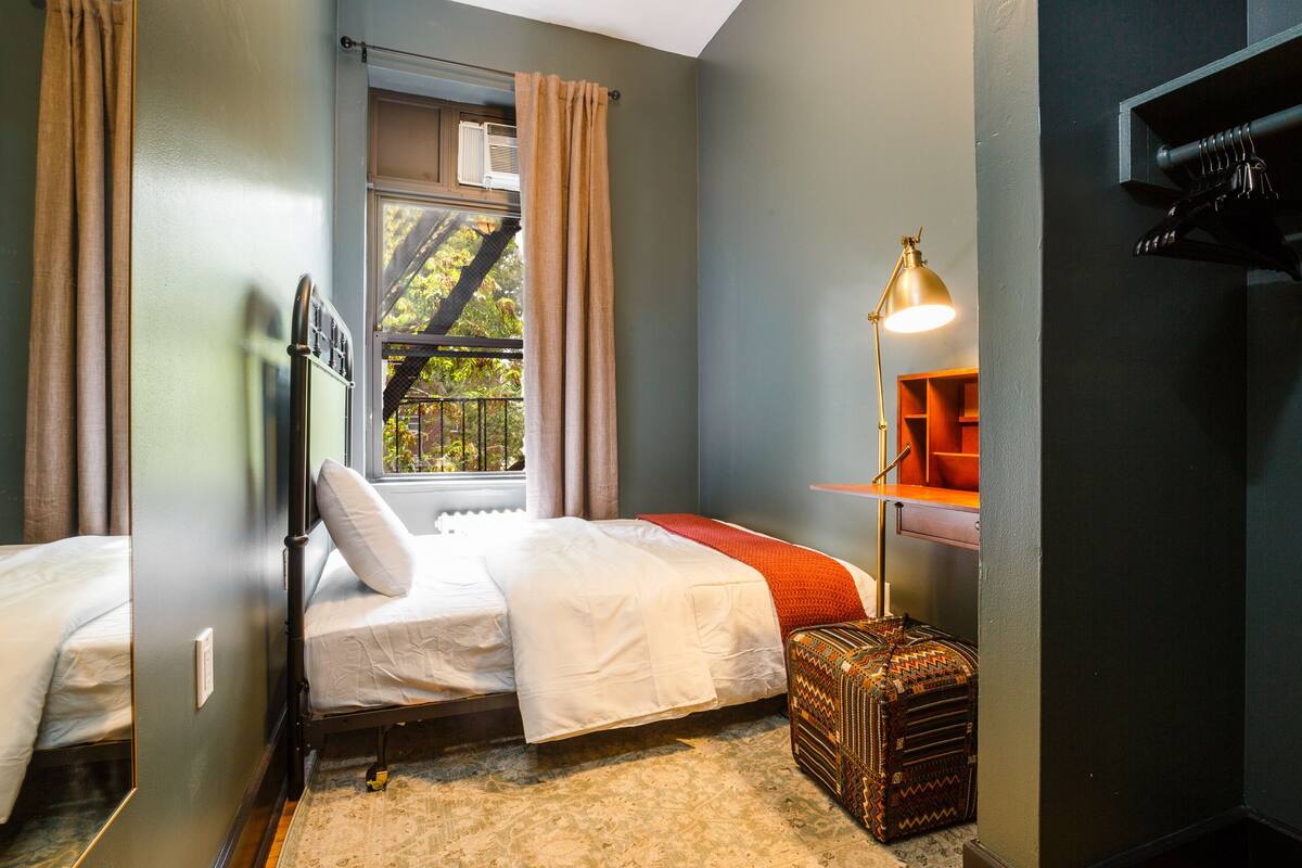 A bedroom with two beds, a window with curtains, a bedside lamp, a small table, and a suitcase.