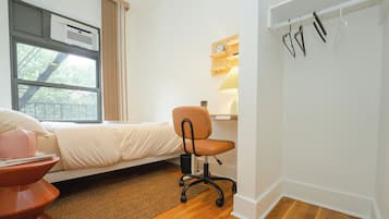 A bedroom with a bed, a desk, a chair, a window, and a wall-mounted shelf.