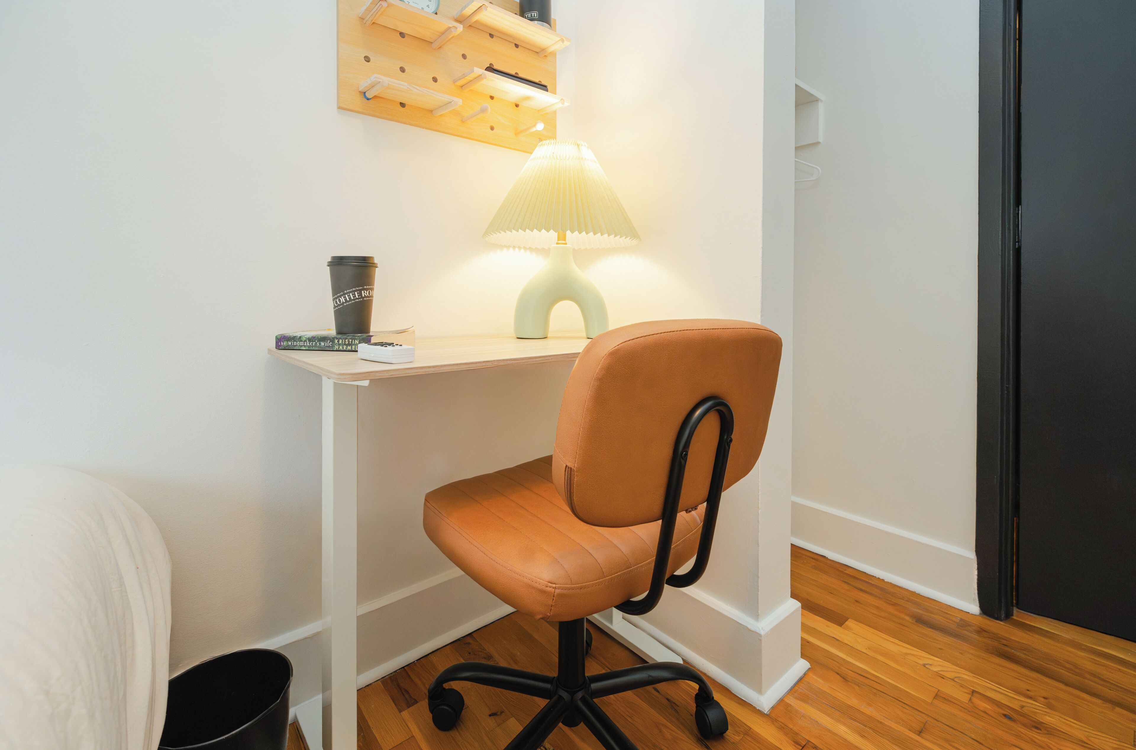 Una habitación pequeña y ordenada, con un escritorio de madera, una silla acolchada, una lámpara y una taza de café.