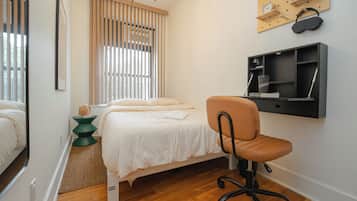 A bedroom with a bed, a desk with a chair, a window with blinds, and a wall-mounted shelf with a clock and headphones.