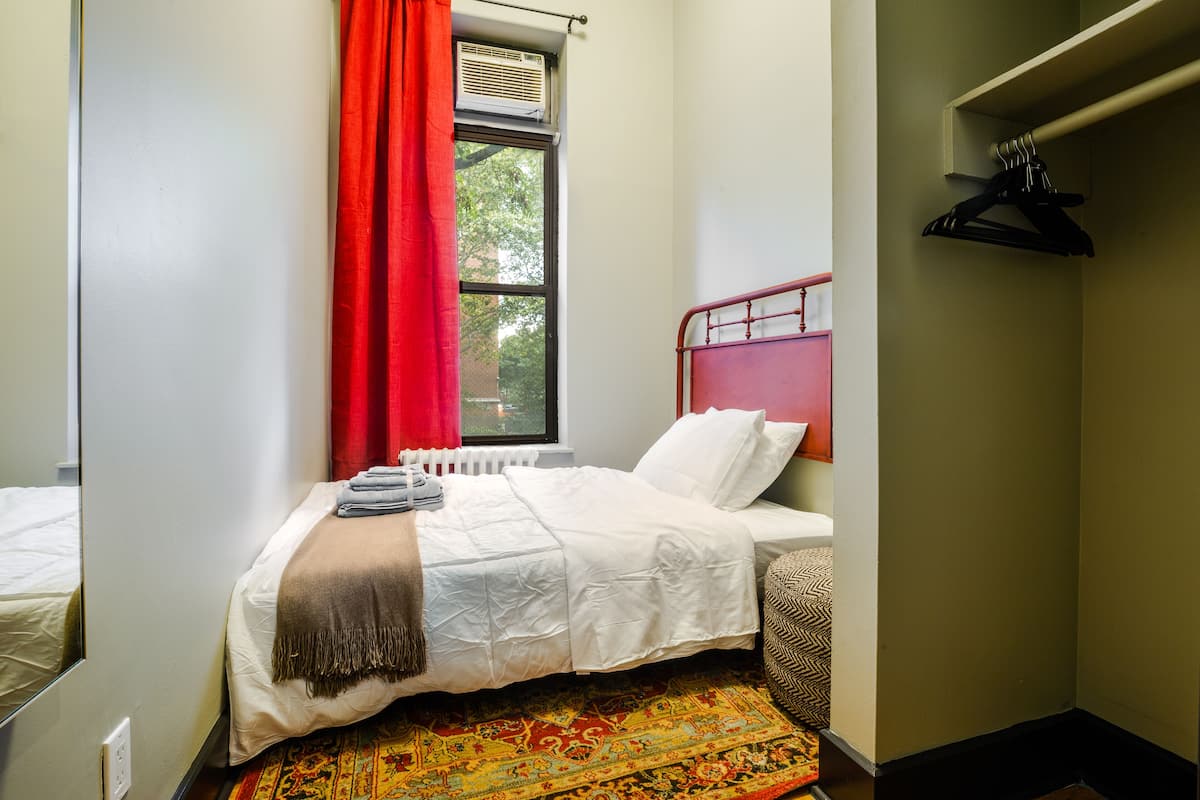 A neatly made bed with a white comforter and a brown throw blanket, a red curtain, and a radiator above the window.