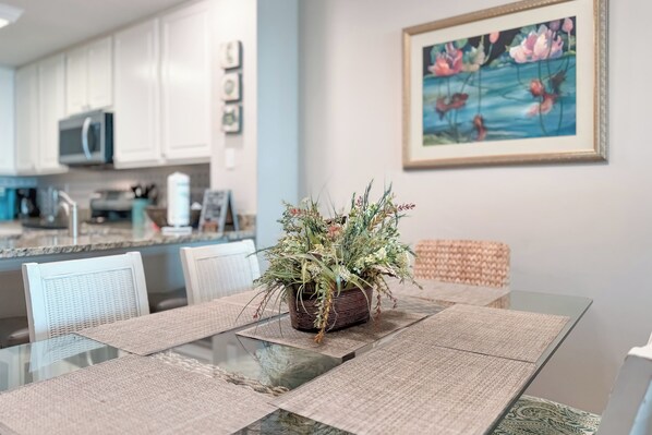 Un comedor con una mesa de vidrio, individuales tejidos y un centro de mesa con planta en maceta. Se ven sillas blancas y una silla tejida.