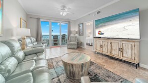 Een woonkamer met een grote televisie waarop een strandscène te zien is, een bank, een gekleurd vloerkleed en een plafondventilator.