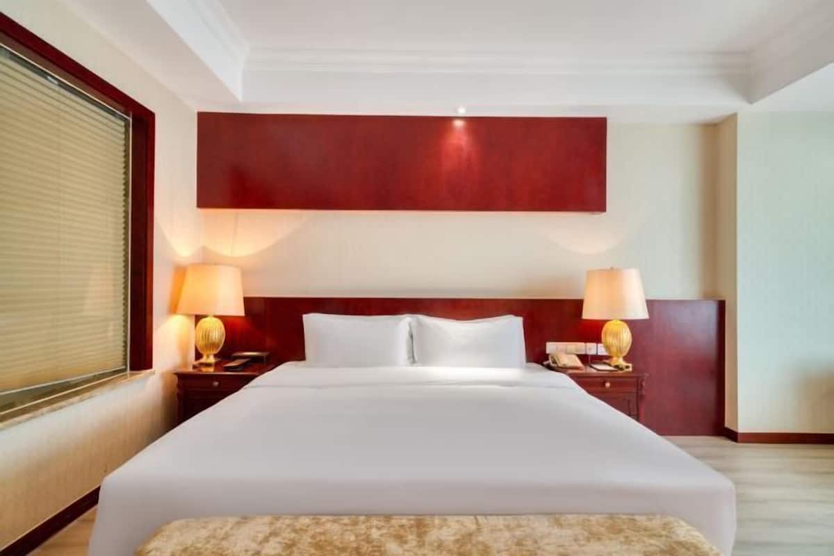 Une chambre d’hôtel avec un grand lit, deux tables de chevet avec des lampes, un mur d’accent rouge et une fenêtre avec des stores.