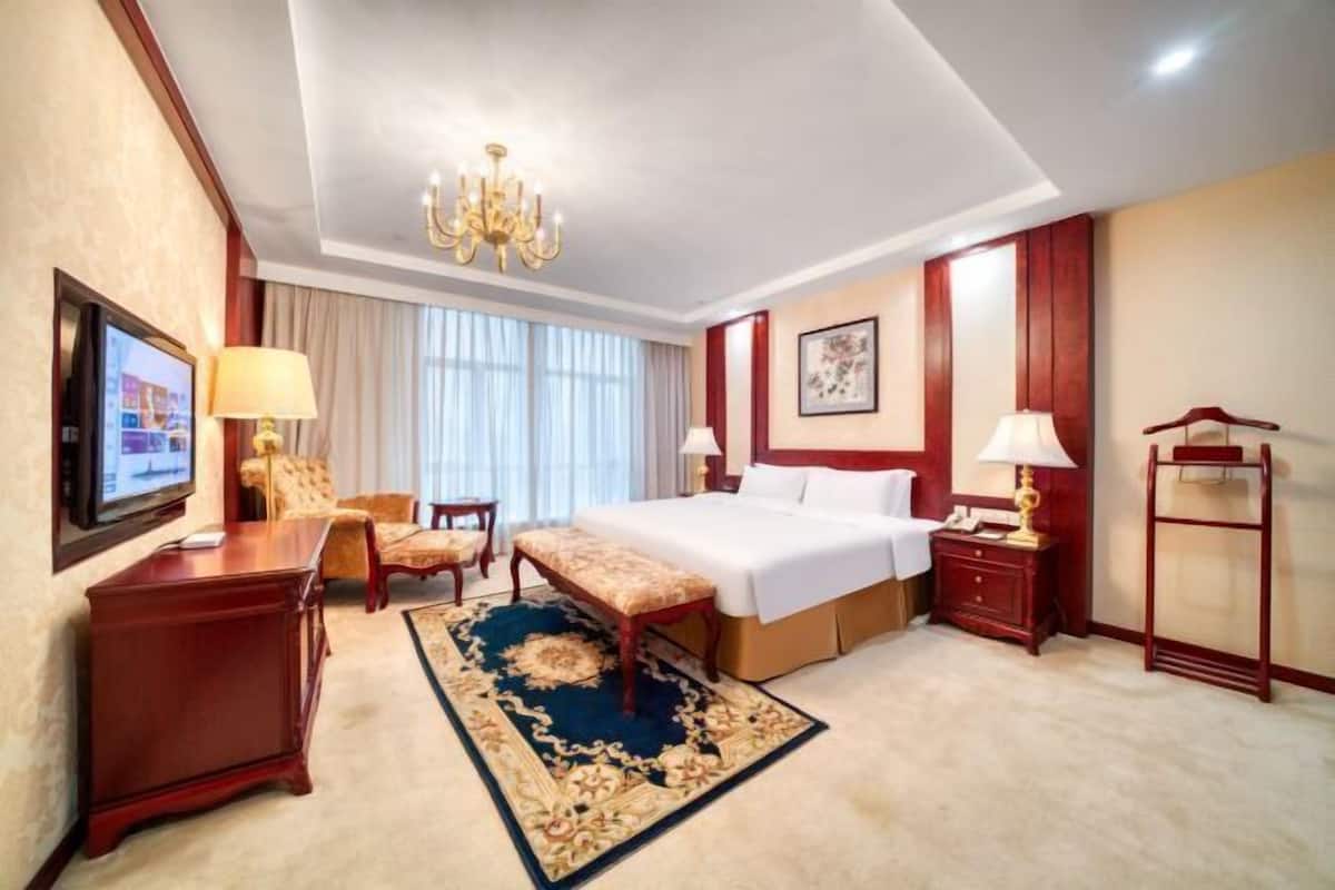 Une chambre d’hôtel avec un grand lit, un bureau, une chaise, un banc, une télévision et un lustre.