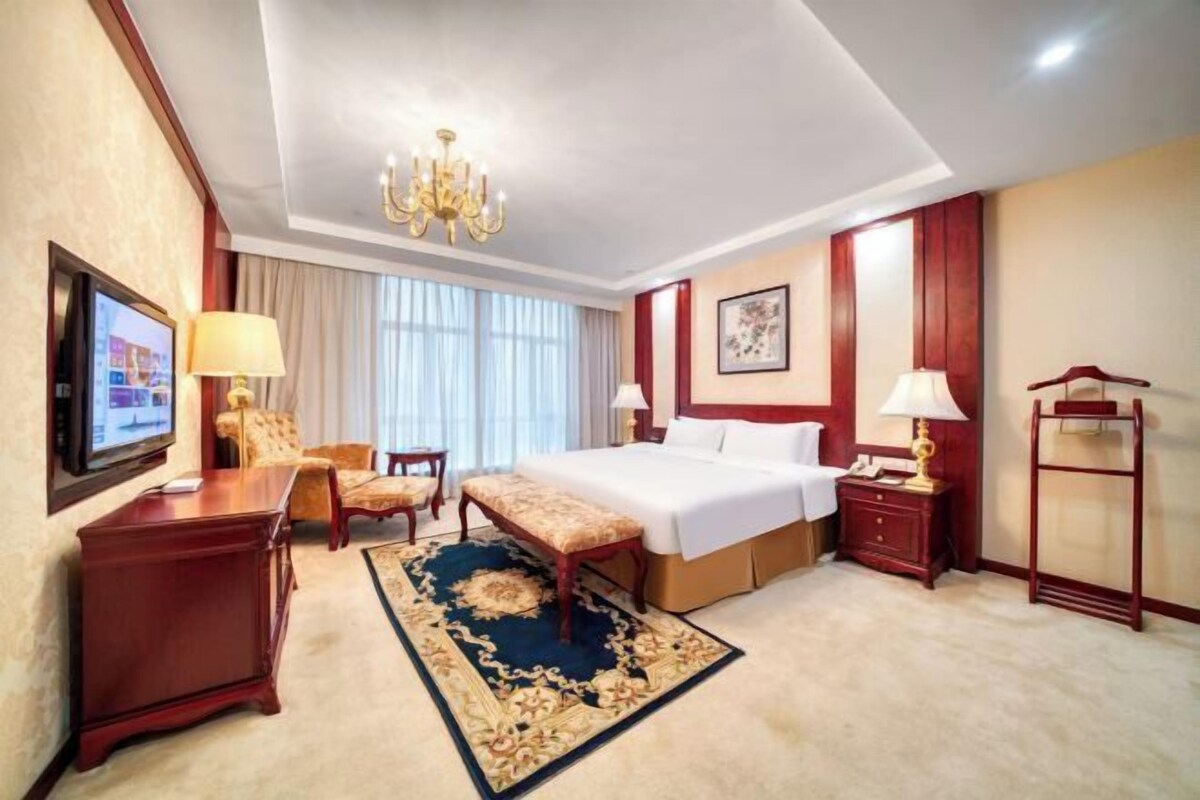 Een hotelkamer met een bed, een bureau, een stoel, een bank, een televisie, een kroonluchter en een raam met gordijnen.