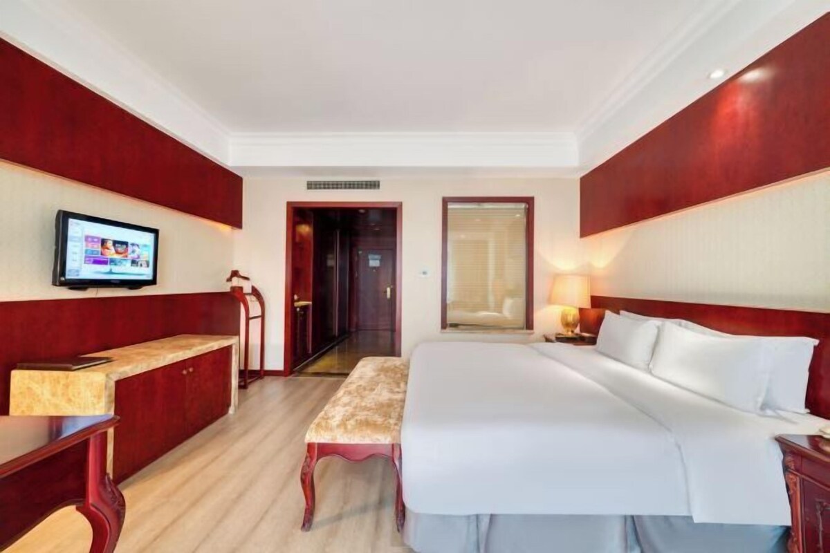 Een hotelkamer met een groot bed, een bureau met een computer, een bankje en een televisie aan de muur.
