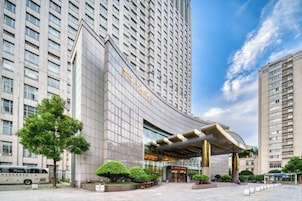 Un hotel moderno con fachada de vidrio curvo, una entrada destacada y un espacio exterior bien cuidado con árboles y cielo despejado.