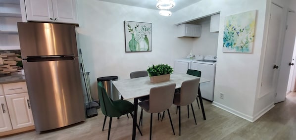 A modern kitchen with a stainless steel refrigerator, a dining table with chairs, and a potted plant. There are two wall-mounted washing machines and a ceiling light fixture.