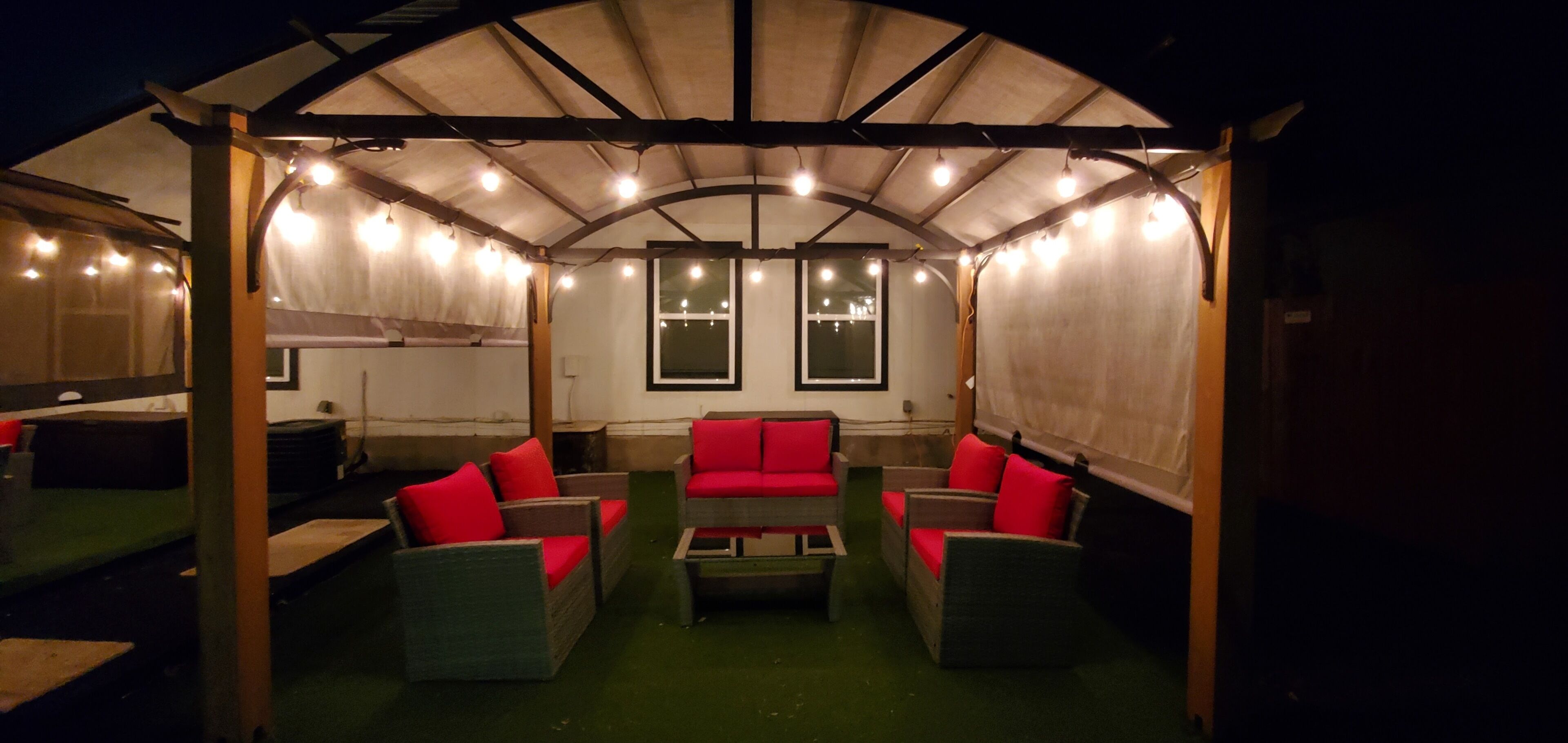 Un espace extérieur aménagé avec des meubles en osier et des coussins rouges, protégé par un toit et illuminé de guirlandes lumineuses.