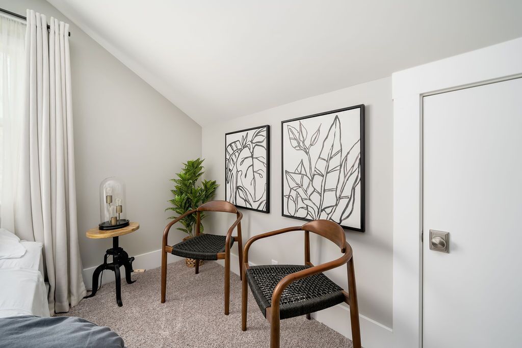 Un dormitorio con una cama, dos sillas, una mesita con una lámpara y dos cuadros enmarcados en la pared.