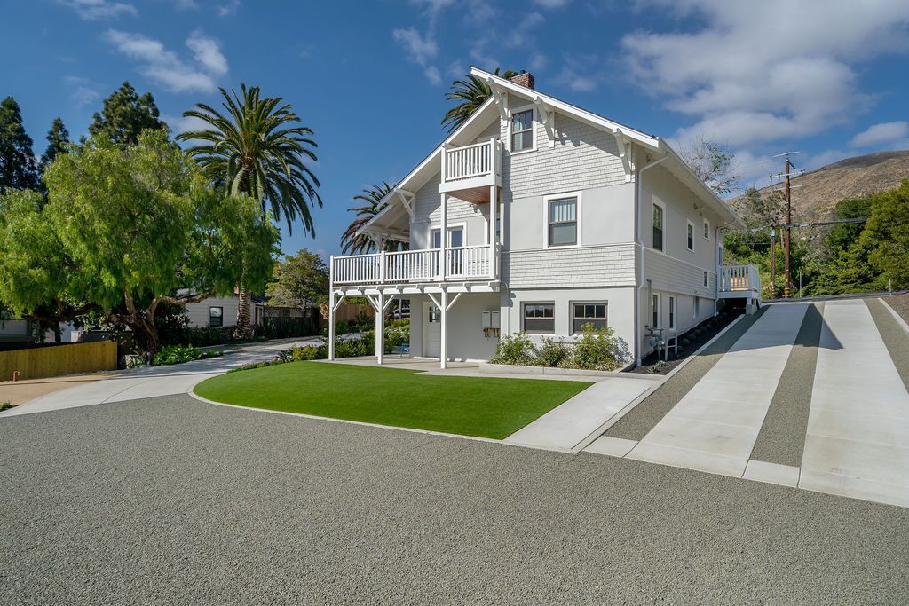 A two-story white house with a balcony, surrounded by greenery and a clear sky.