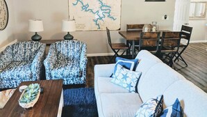 Een woonkamer met een bank, twee fauteuils met een patroon, een salontafel met een plant, een kaart aan de muur, en een eethoek met een tafel en stoelen.