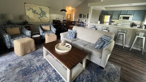 Een woonkamer met een bank, fauteuils, een salontafel en een eethoek met stoelen en een tafel. Achtergrond toont een keuken met een magnetron en barkrukken.