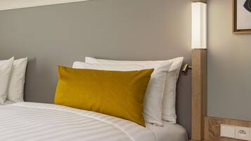 Una cama bien hecha con almohadas blancas y amarillo mostaza, una mesita de noche de madera con interruptor de luz y una pared gris.