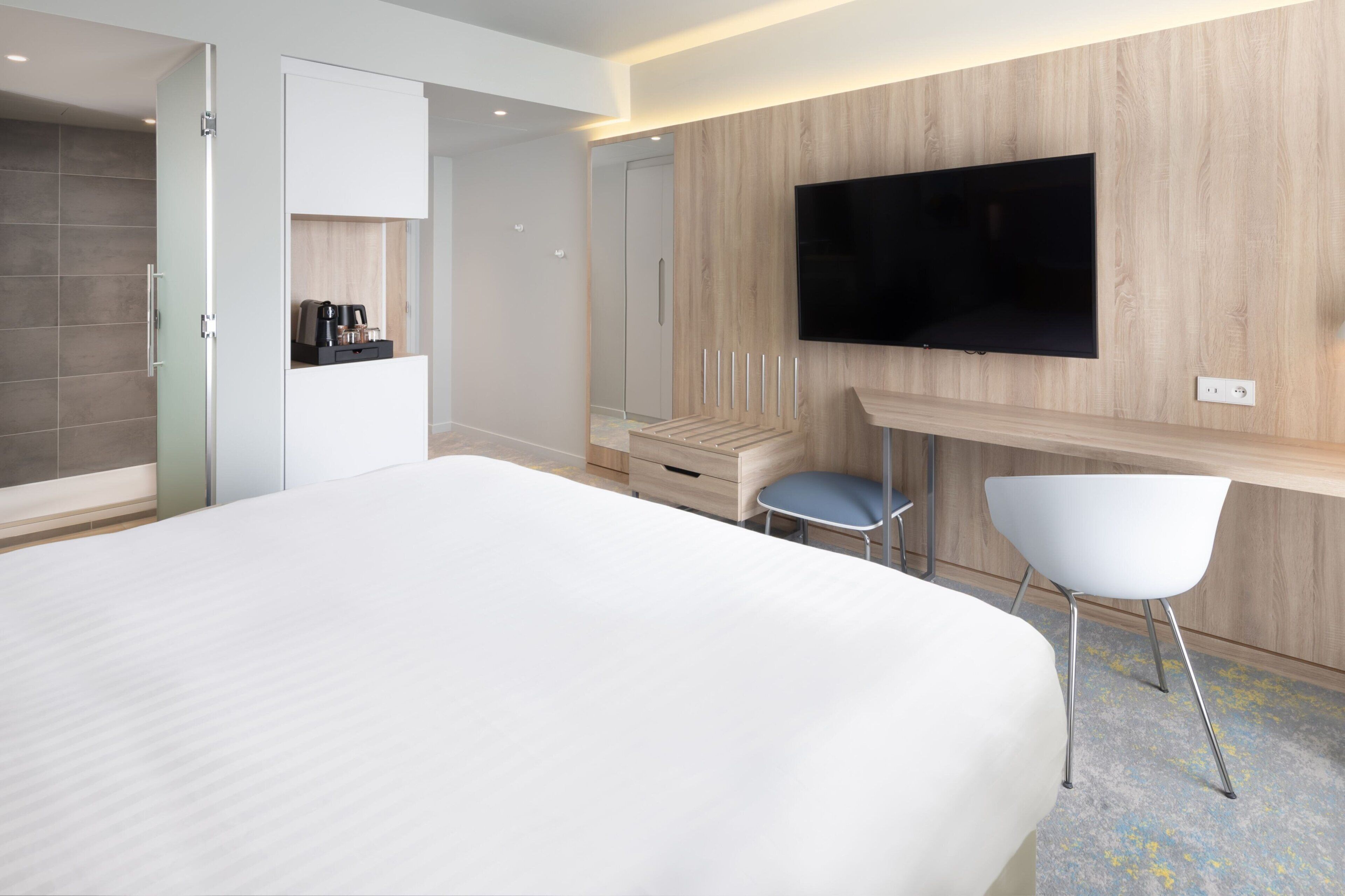 Une chambre d’hôtel moderne dotée d’un grand lit, d’un téléviseur à écran plat fixé au mur, d’un petit coin salon avec un fauteuil et d’un espace dédié à la préparation du café.