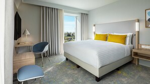 A hotel room with a large bed, a desk with a chair, a lamp, and a view of the outdoors. - Courtyard by Marriott Paris Charles de Gaulle Central Airport (Roissy-en-France)