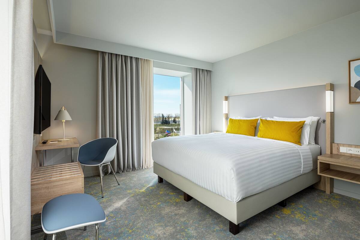 Habitación de hotel con una cama grande, un escritorio con silla, una lámpara y vistas al exterior.
