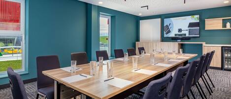 A conference room with a long table, chairs, a television screen, and a window with blinds.