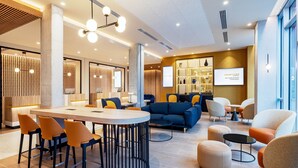 A modern lounge area with a central round table, orange chairs, a blue sofa, and a bar area with shelves. - Courtyard by Marriott Paris Charles de Gaulle Central Airport (Roissy-en-France)