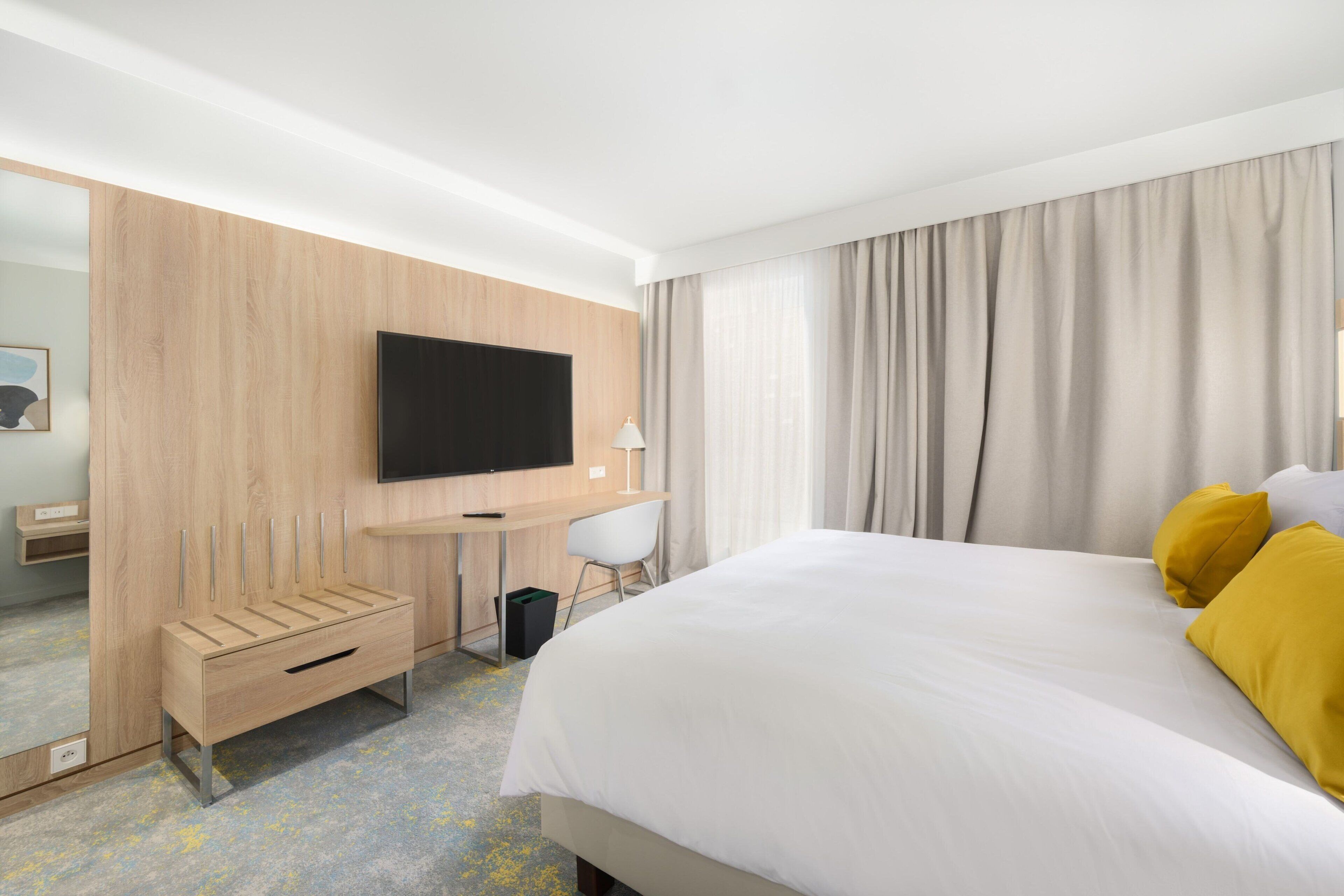 Ein Hotelzimmer mit einem großen Bett, einem Schreibtisch, einem Flachbildfernseher, einem Sessel und einem Holzschrank.