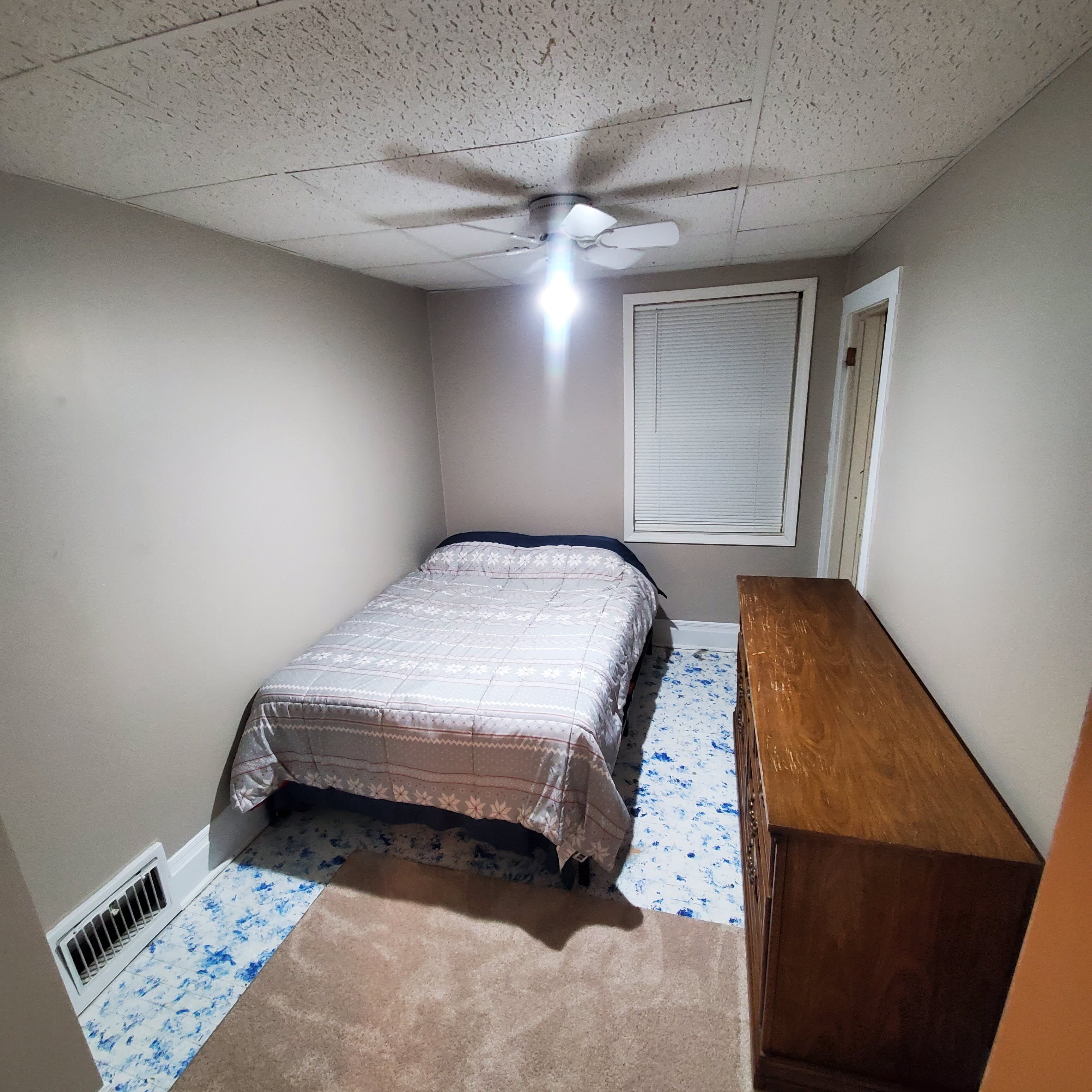 A bedroom with a bed, a wooden dresser, a ceiling fan, a window with blinds, and a vent.