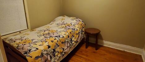 Un dormitorio pequeño con una cama individual, piso de madera y un taburete.
