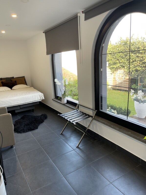 A modern bedroom with a large window, a bed with pillows, a small table with a plant, and a black rug on the floor.
