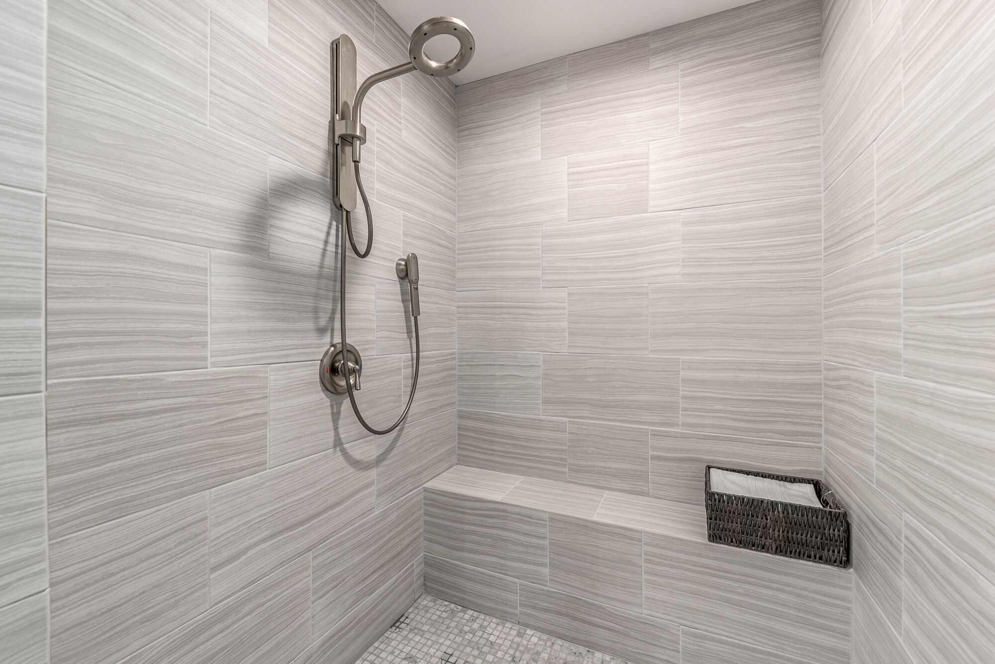 A shower with a handheld showerhead and a wall-mounted faucet, a small bench, and a wicker basket.