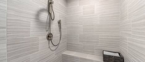 A shower with a handheld showerhead and a wall-mounted faucet, a small bench, and a wicker basket.