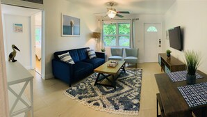 A living room with a blue sofa, a wooden coffee table, and a patterned rug. There is a ceiling fan, a TV mounted on the wall, and a door leading to another room.