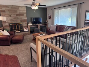 A living room with a stone wall, a ceiling fan, a flat-screen TV, a brown leather sofa, and a wooden coffee table. - Mountain View...walk to attractions & dining (Yelm)
