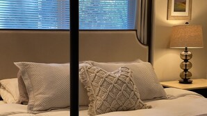 Ein ordentlich bezogenes Bett mit einem gemusterten Zierkissen und einem schlichten Kopfkissen, einer Nachttischlampe und einem Fenster mit Jalousien.