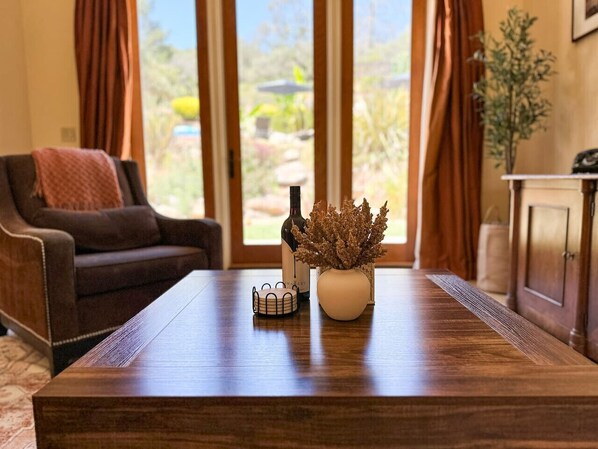 A wooden table with a bottle and a vase of dried plants, a small decorative item, and a view of a garden through large windows.