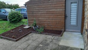 Ein Holzhaus mit einer grauen Tür, ein kleiner Garten mit einem Kaktus und einer gepflasterten Fläche.