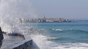 Eine Küstenansicht mit brechenden Wellen an einer Betonbarriere, einer entfernten Stadtansicht und blauem Himmel.