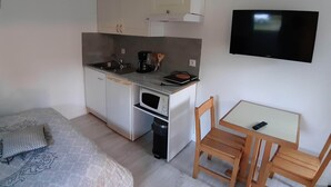 Eine kompakte Küchenzeile mit Mikrowelle, Kaffeemaschine und Spüle. Ein kleiner Tisch mit Stuhl, ein an der Wand montierter Fernseher und ein Bett mit gemustertem Überwurf sind vorhanden.
